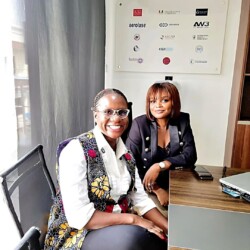 Two Professional Women Collaborating In A Modern Office, Smiling And Engaged During A Productive Work Session. Two Professional Women Collaborating In A Modern Office With Natural Light And Company Branding.